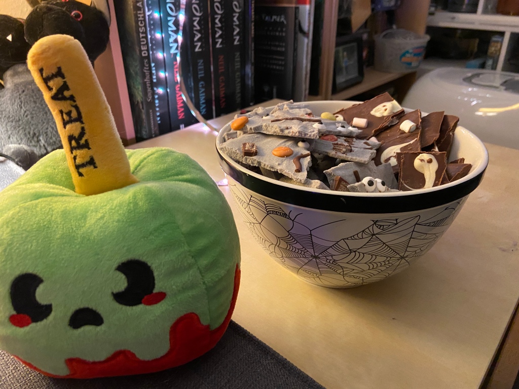 Links vorn im Bild steht ein Plüsch-Apfel in grün und rot mit einem Gesicht und der Aufschrift TREAT. Rechts daneben steht eine Schale mit zweierlei Schokolade: Lila mit Zuckeraugen und Vollmilch mit aufgemalten Gespenstern. 