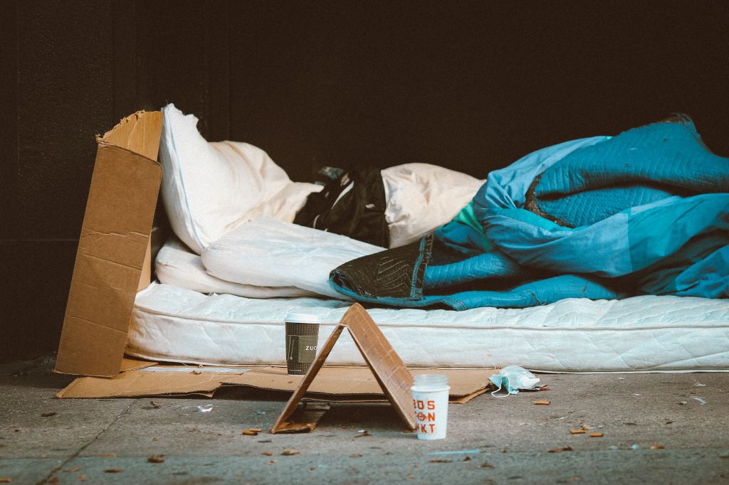 Ein Obdachlosenlager. Kartons, eine Matratze, mehrere abgenutzte Decken und Kissen. Auf dem Boden davor ein Schild aus Pappe (nicht lesbar) und ein Becher für Mpnzen. Der Boden ist mit Unrat bedeckt.