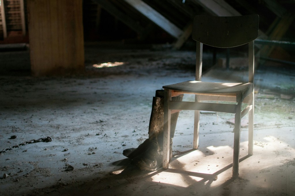 In einem verlassenen Raum steht ein einsamer Holstuhl neben einem abgetragenen Paar Stiefeln. Von schräg rechts fällt Sonnenlicht in den Raum. Der Boden ist schmutzig, eine Kette liegt herum. Der Raum wirkt verlassen. Möglicherweise handelt es sich um einen Dachboden.